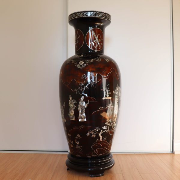 Dark brown traditional Vietnamese lacquer and mother-of-pearl vase. This side of the vase is depicting a notable man watching music players. Next to him, another man stands near an archway, possibly symbolizing an entrance to a garden or a ceremonial site. The background maintains the serene landscape with mountains and trees.